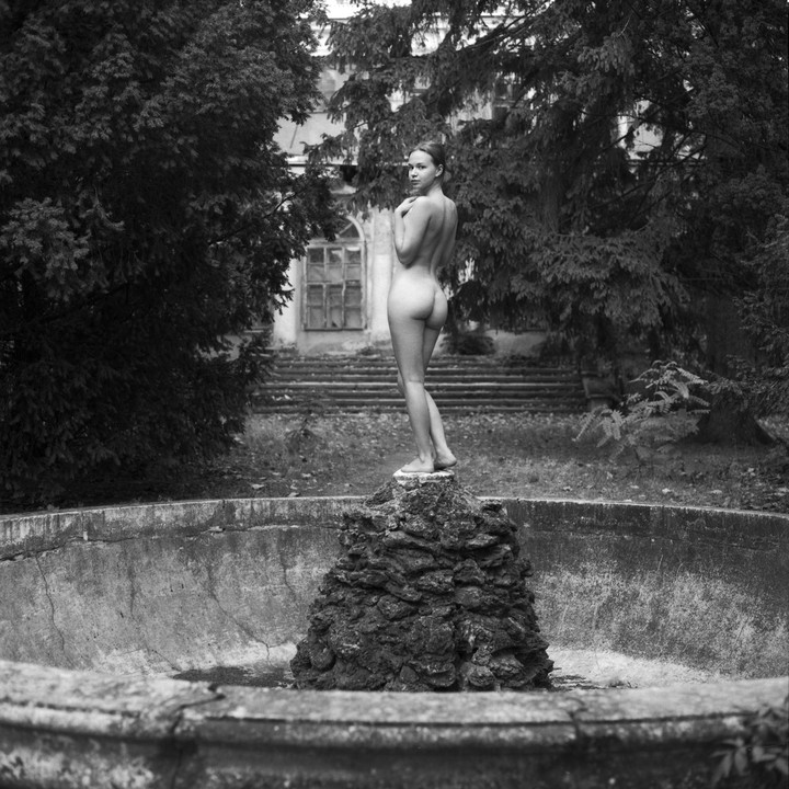 A nude woman stands on a rock formation in a dry fountain, looking over her shoulder at the viewer, with a building and trees in the background.