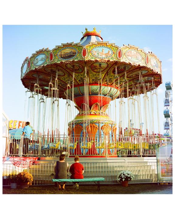 Two people sit on a bench in front of a colorful swing ride at a fair.