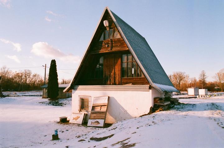 A-frame house with a tall, narrow tree next to it stands in a snowy field under a blue sky.