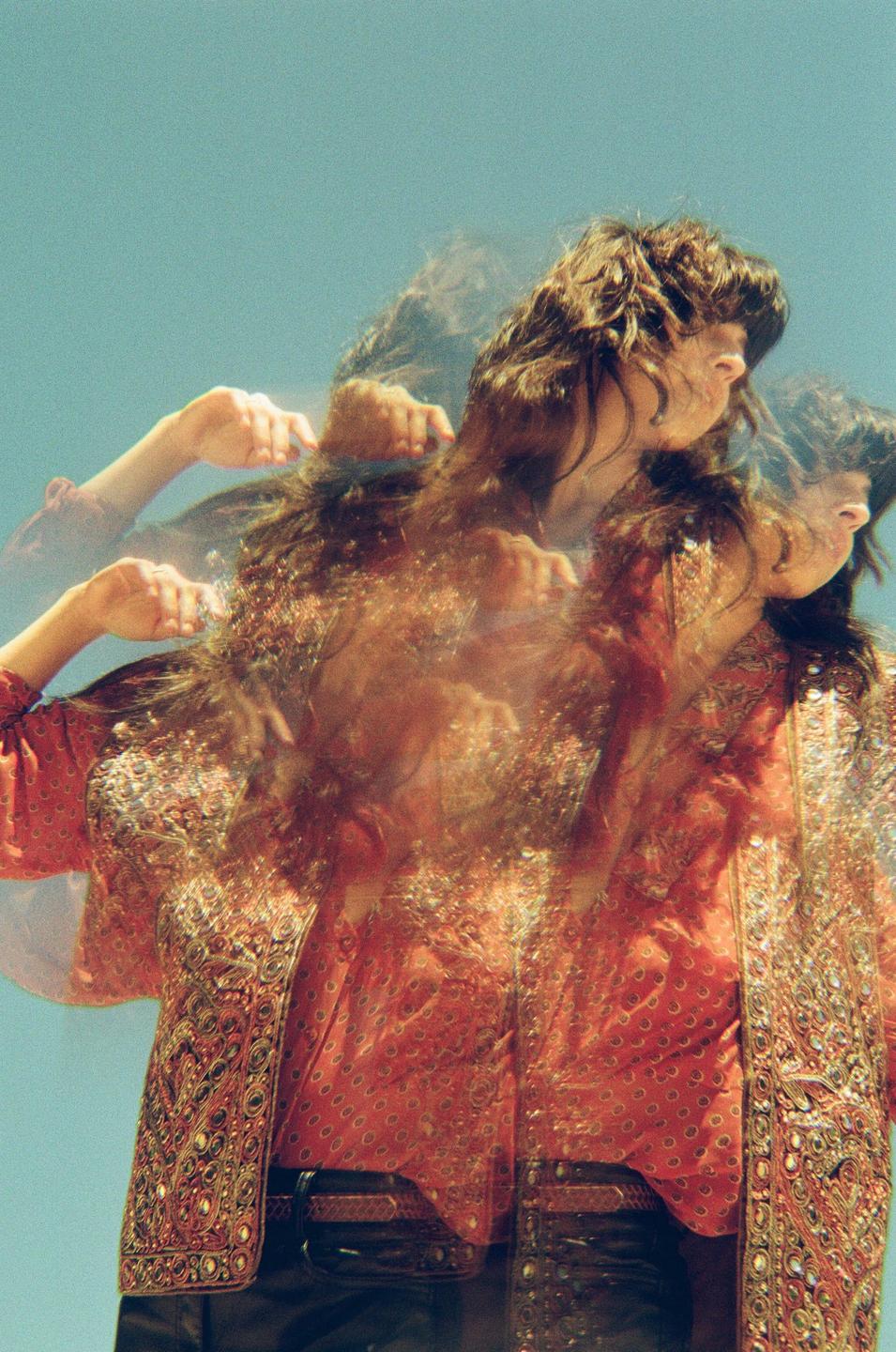 A woman with long hair and a patterned jacket is captured with a multiple exposure effect against a clear sky.