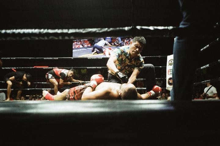 A man in a floral shirt attends to a fighter lying on the canvas in a boxing ring.