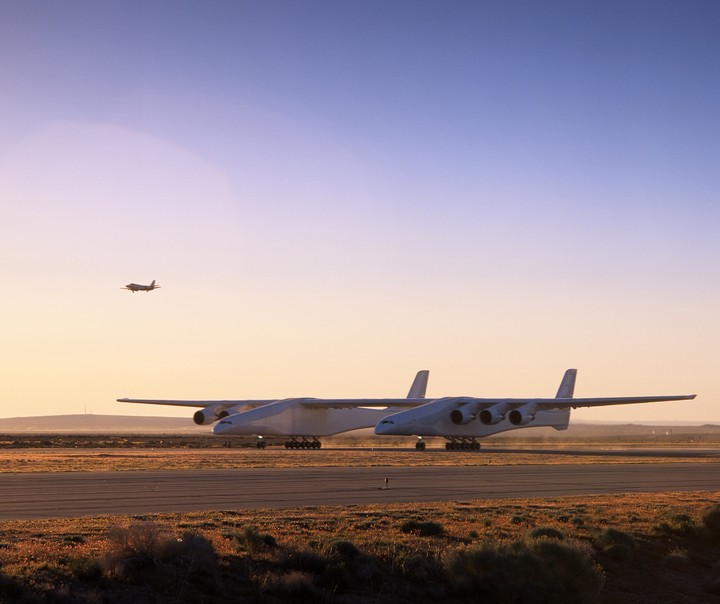 Two large white aircraft are on a runway with a smaller plane flying in the distance under a gradient sky.