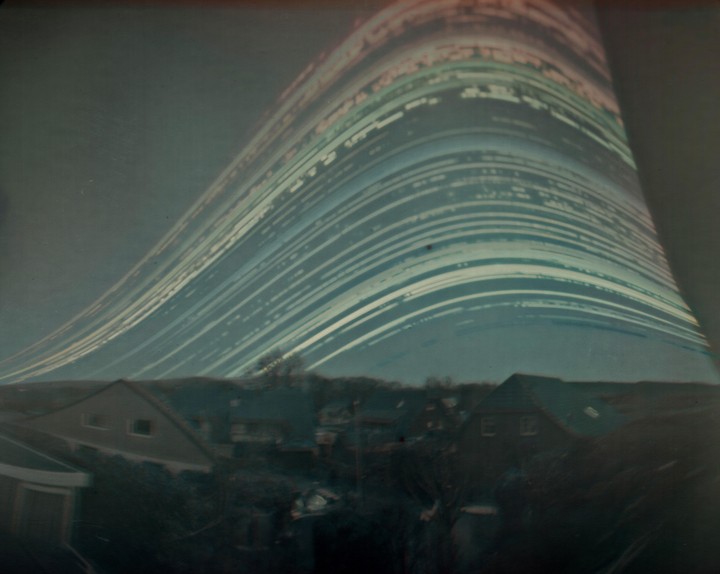 A long exposure photograph captures star trails curving over a small town with houses and trees.