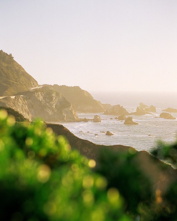 A winding road follows a rugged coastline with sea stacks in the water under a hazy sky.