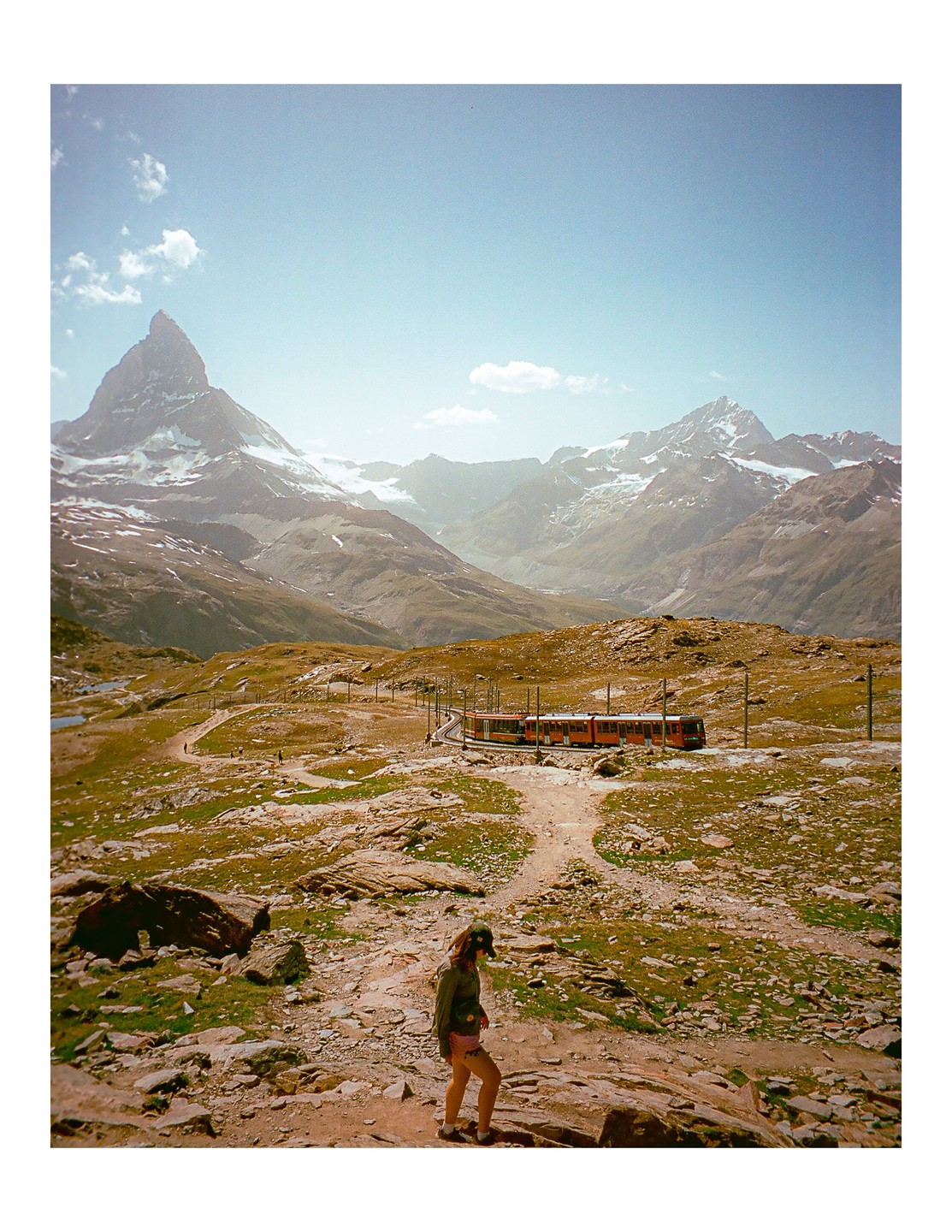 A woman walks on a rocky path with a train on tracks and snow-capped mountains in the background.