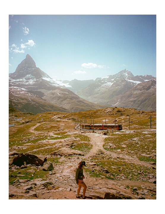 A woman walks on a rocky path with a train on tracks and snow-capped mountains in the background.