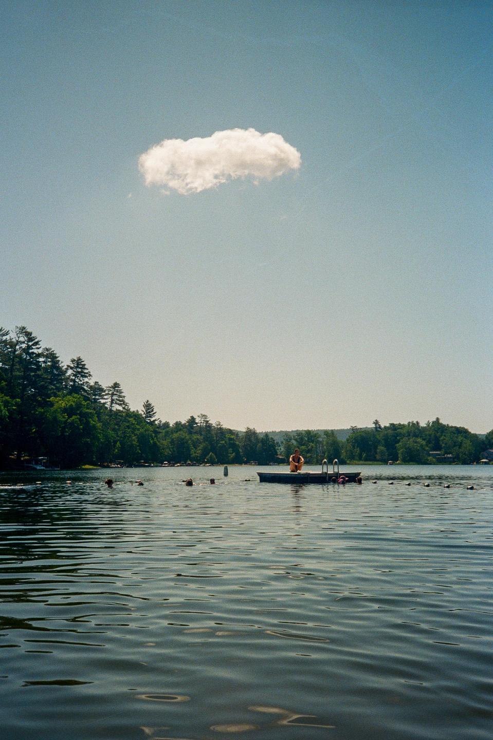 A person sits on a floating dock in a lake with trees on the far shore under a clear sky with a single cloud.