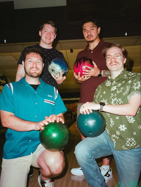 Four men pose with bowling balls at a bowling alley.