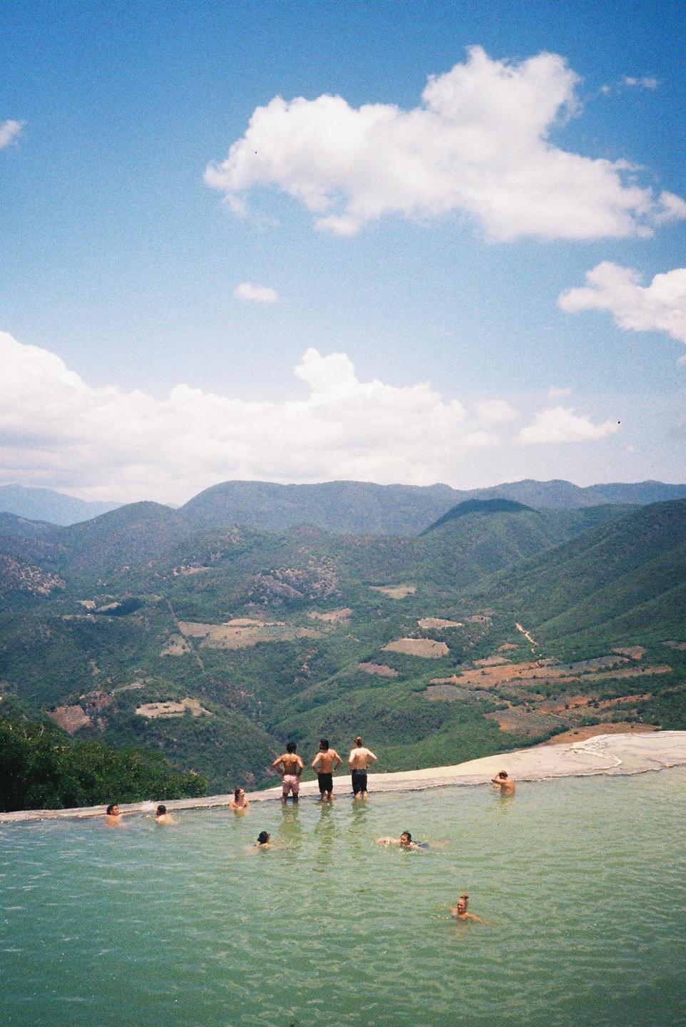 People swim in a natural pool overlooking a mountainous landscape.