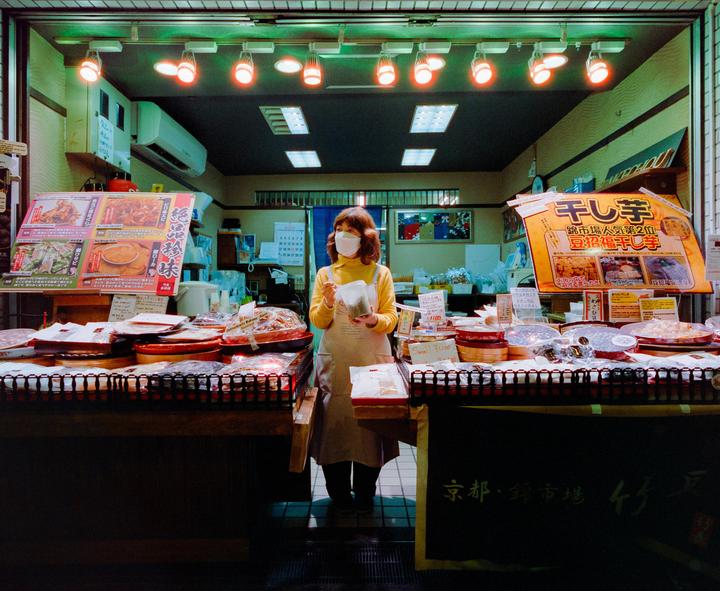 A woman wearing a face mask and apron stands behind a counter filled with food items in a market stall.