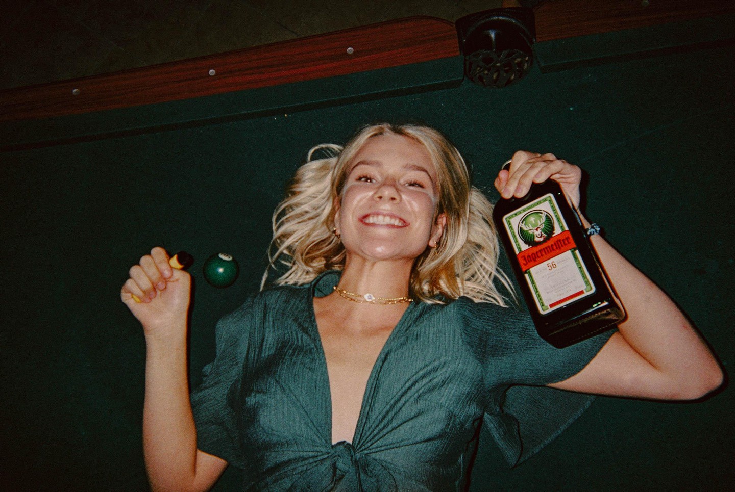 A smiling woman with pigtails lies on a pool table holding a bottle of Jagermeister and a pool cue chalk.