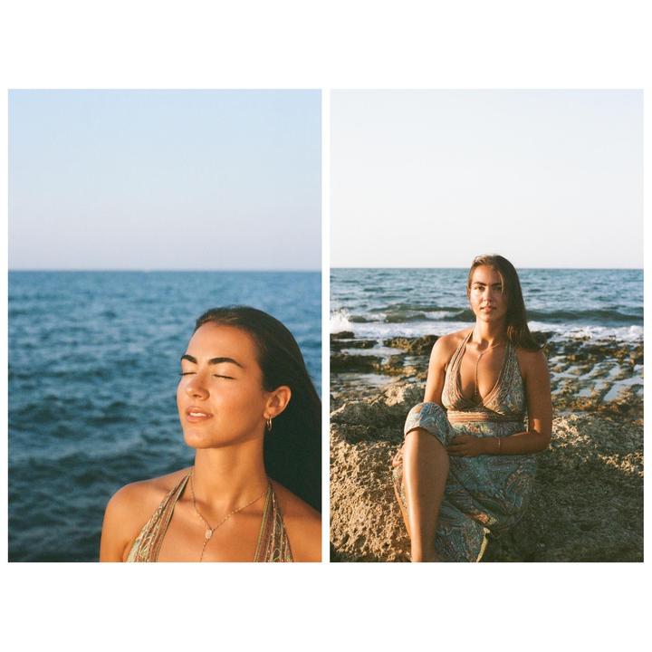 A woman stands with her eyes closed and sits on rocks by the ocean.