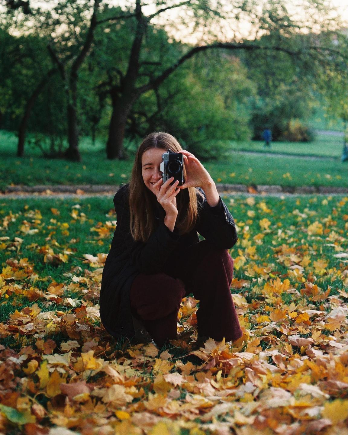 A woman crouches in a park covered in autumn leaves, holding a camera to her face and winking.
