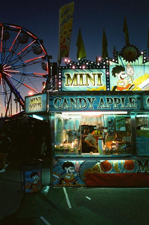 A candy apple stand and a ferris wheel are lit up at a local fair at night.