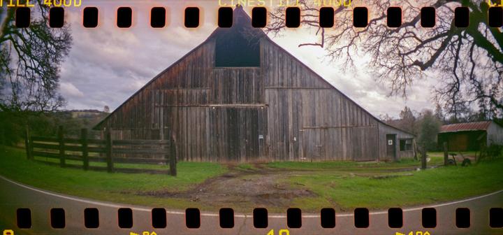 A large wooden barn with a smaller shed next to it is visible from a road with a wooden fence and trees under a cloudy sky.