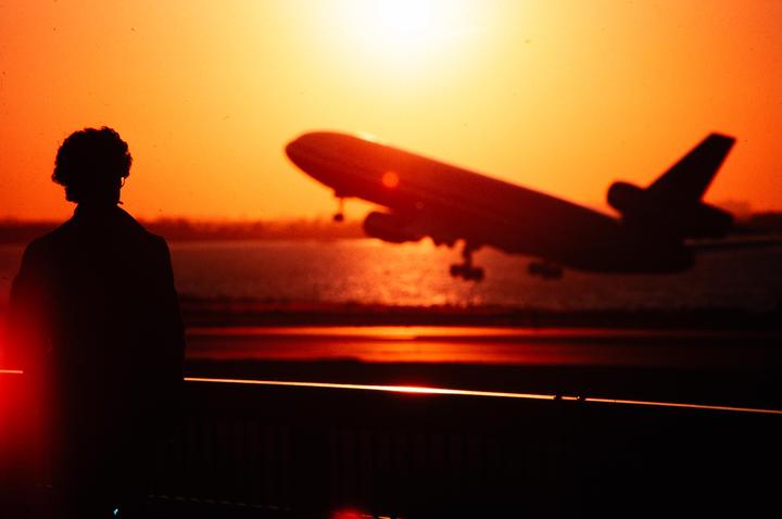 A person watches a plane take off against a bright orange sunset over water.