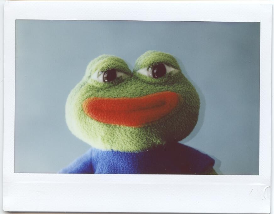 A green frog plush toy with large eyes and an orange mouth is centered against a light blue background.