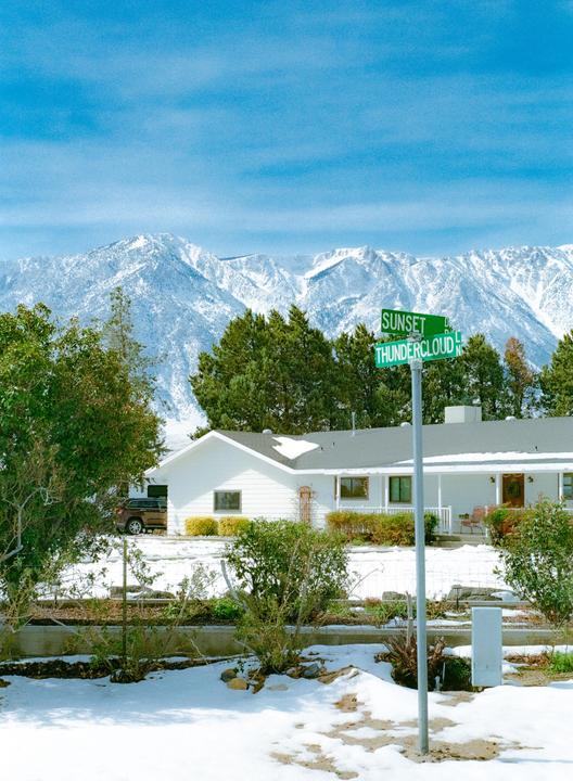 A street sign for Sunset and Thundercloud is in front of a white house with snow-capped mountains in the background.