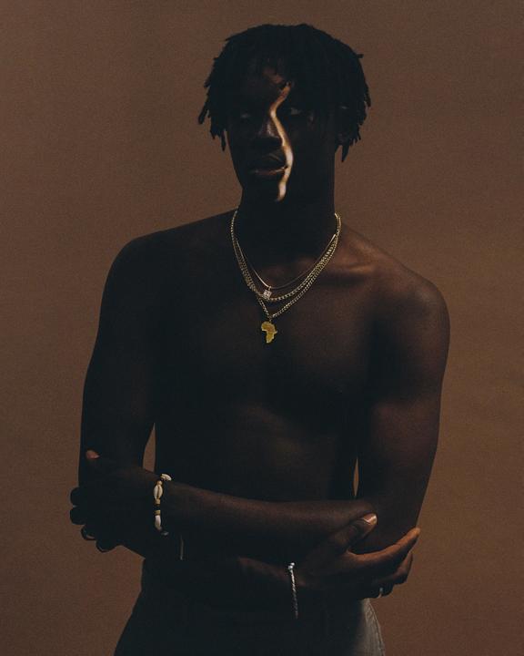 A man with dreadlocks and necklaces stands shirtless with his arms crossed, with a strip of light across his face.