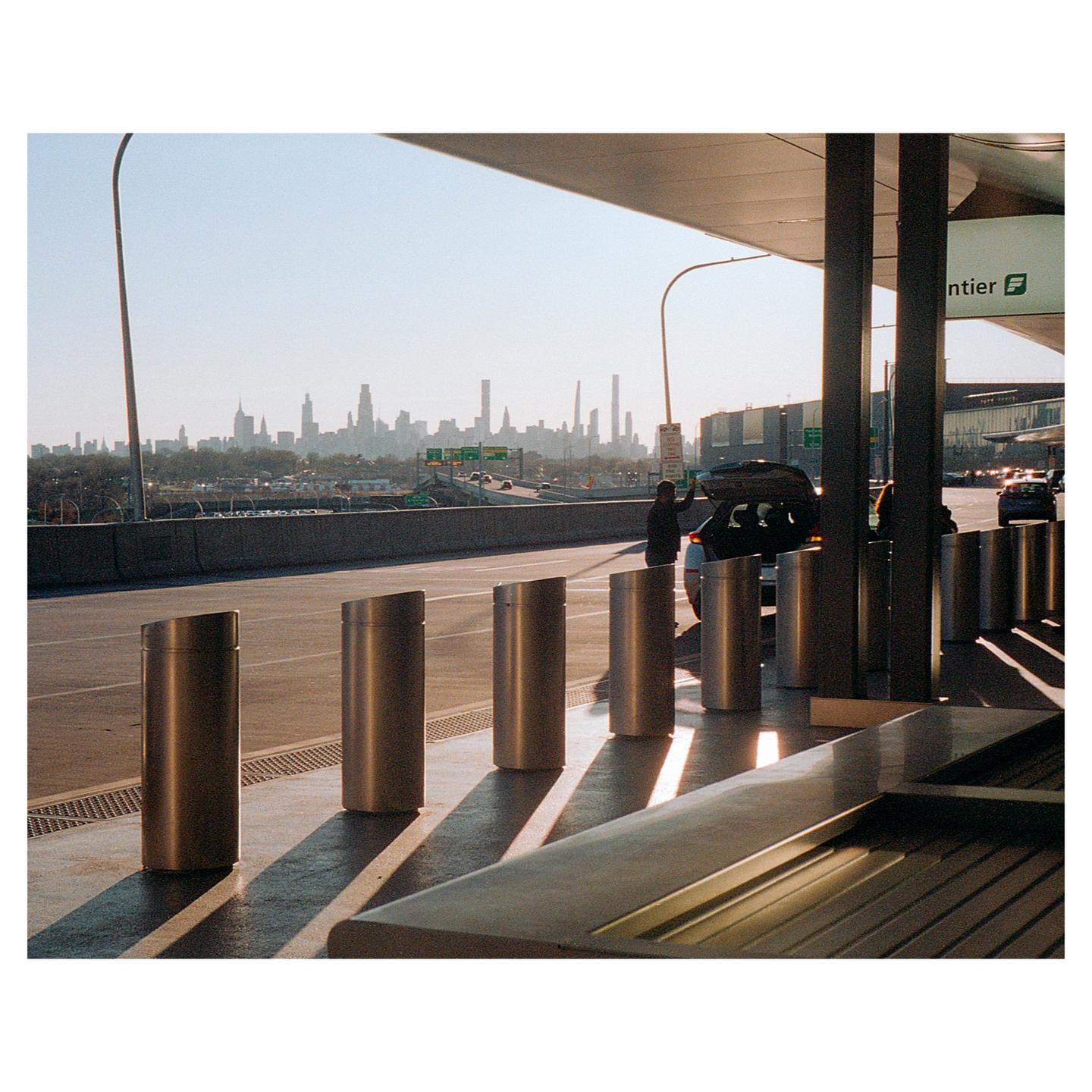 A person stands by a car with an open trunk on a road with a city skyline in the background.