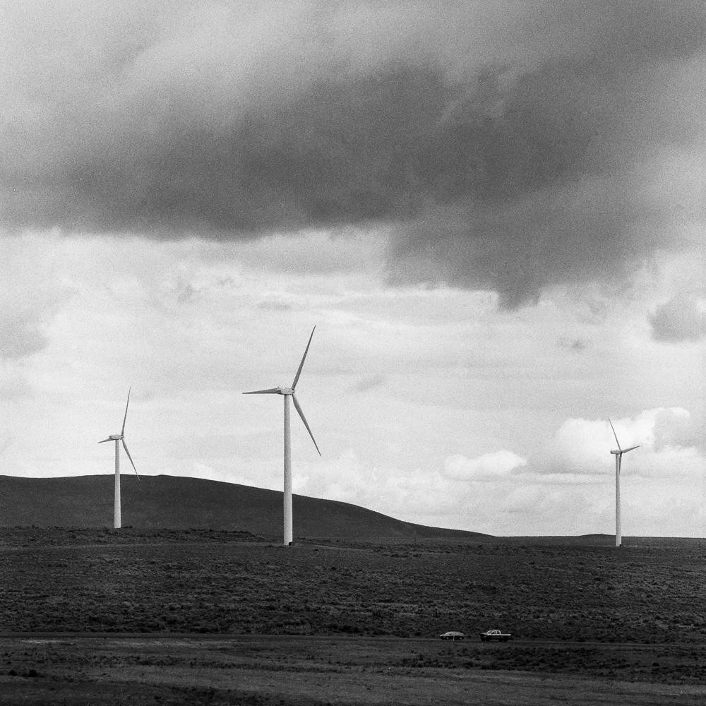 Three wind turbines stand on a hill under a cloudy sky, with two cars visible in the distance.