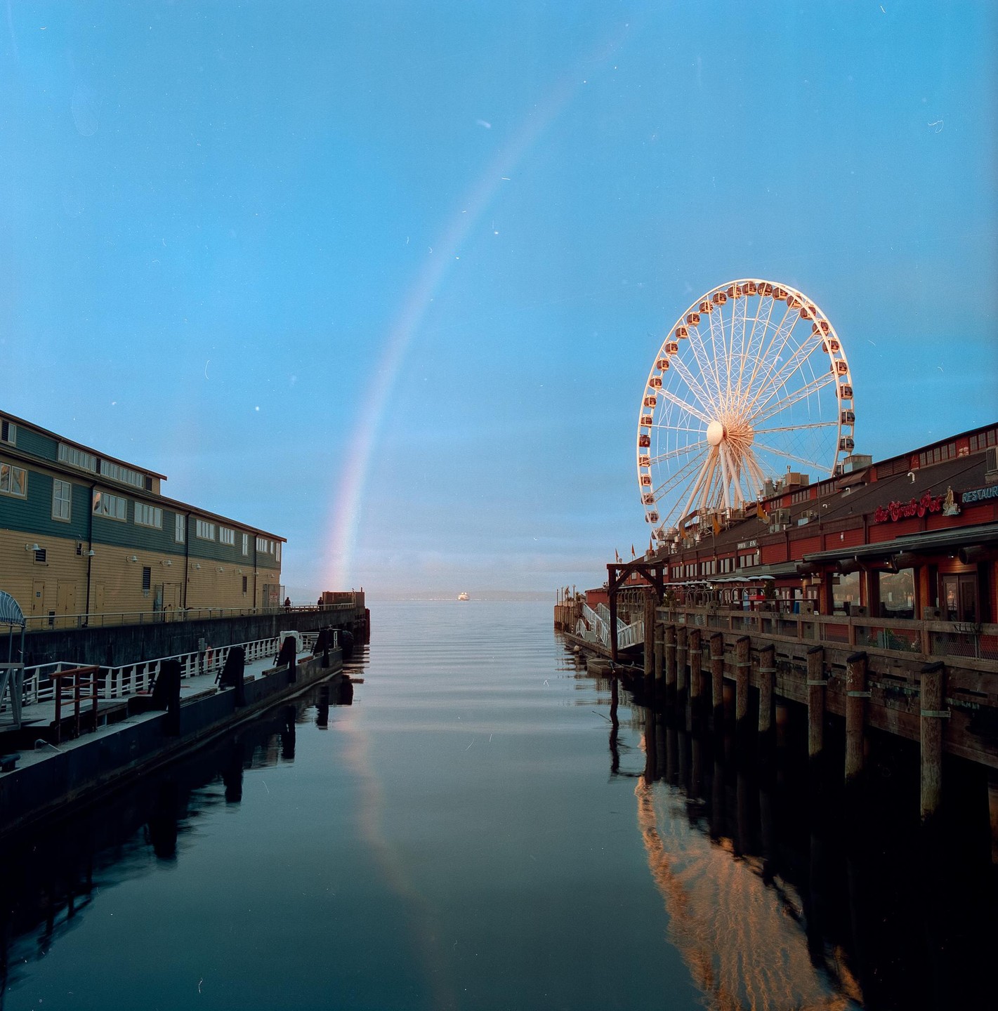 A rainbow arches over a body of water with a Ferris wheel and buildings on either side.