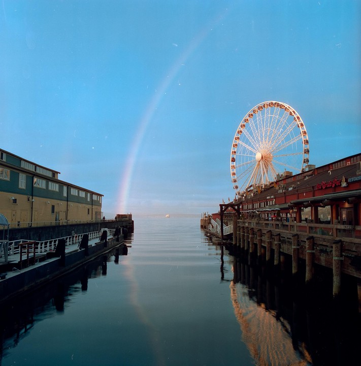 A rainbow arches over a body of water with a Ferris wheel and buildings on either side.