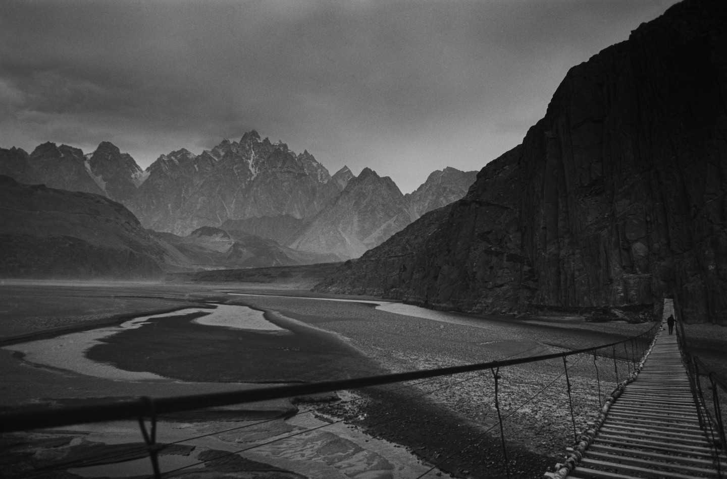 A person walks across a suspension bridge over a river with mountains in the background.