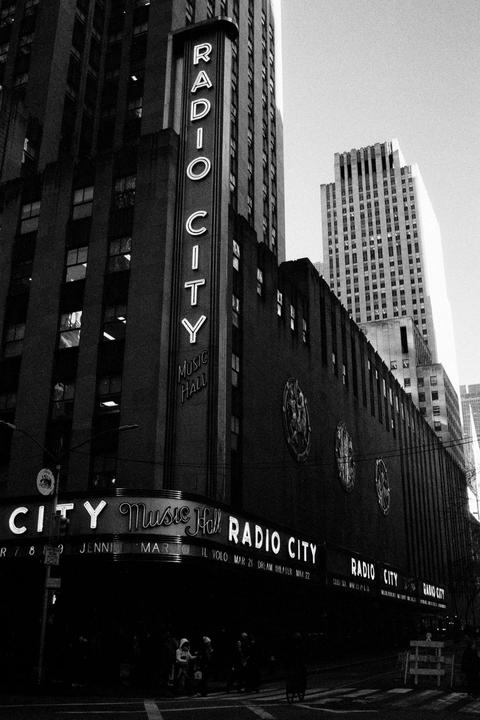 A black and white street view shows the Radio City Music Hall building with its illuminated signs and surrounding skyscrapers.