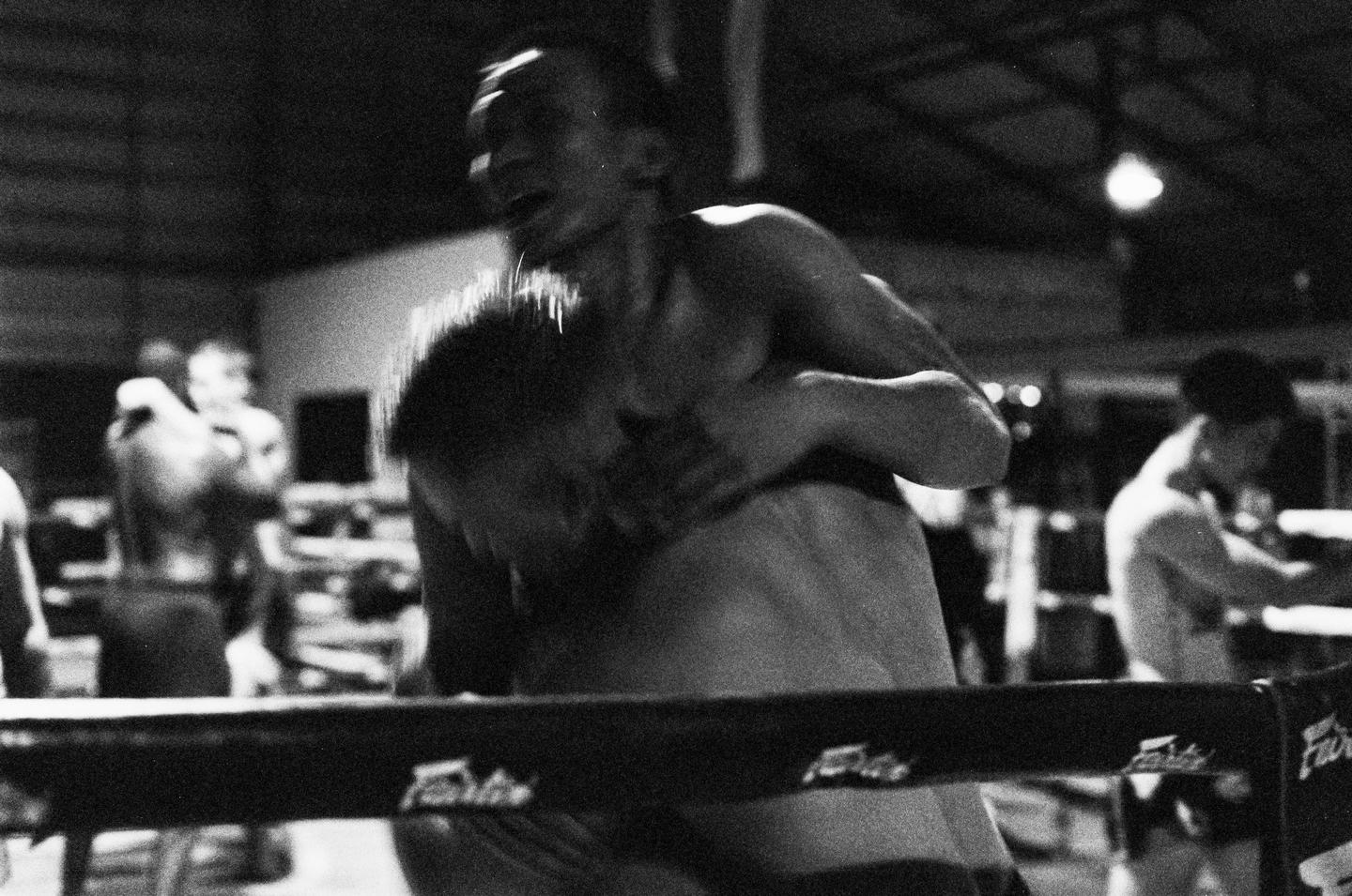 Two men are grappling in a boxing ring with other men visible in the background.