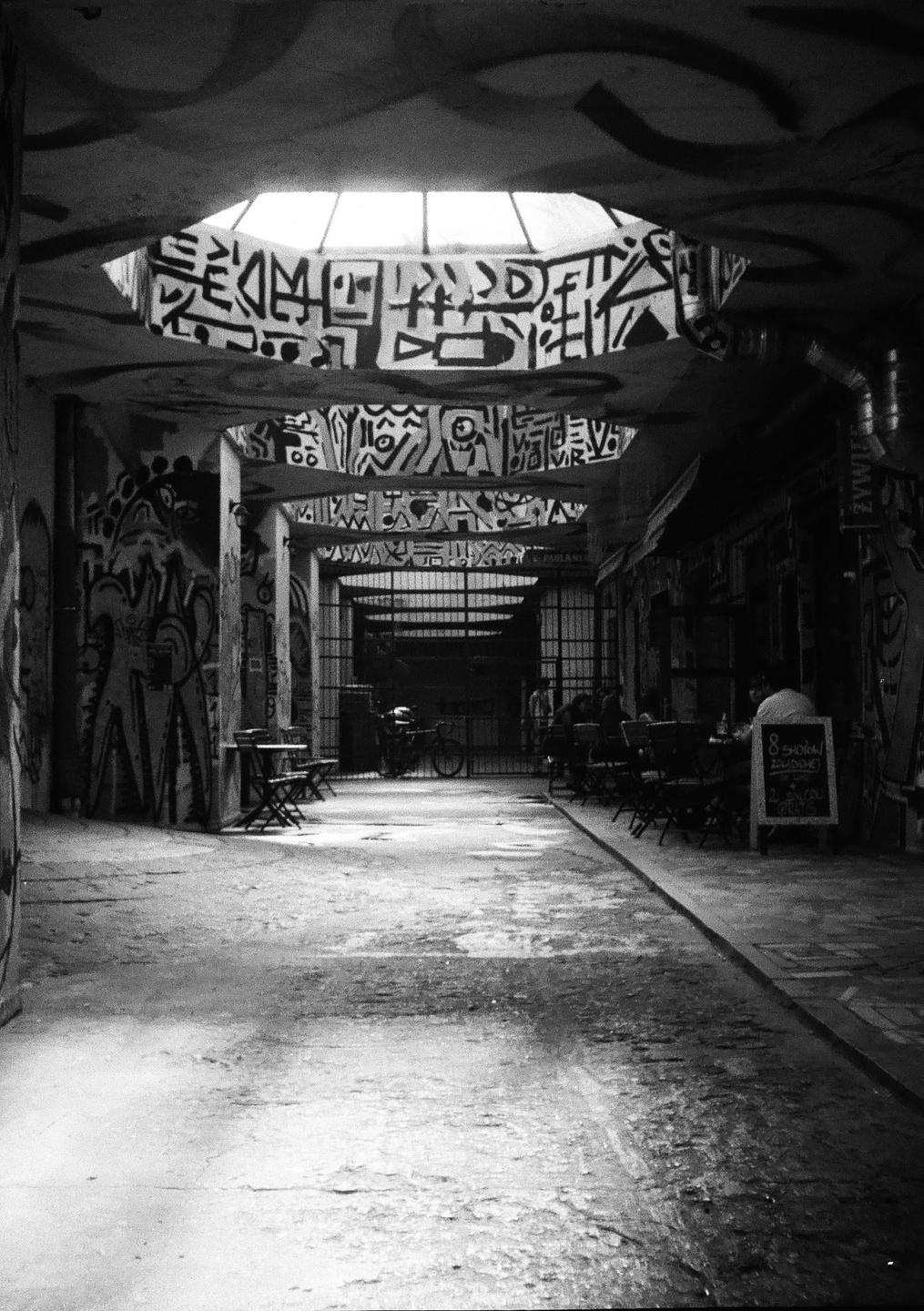 A long, narrow, graffiti-covered corridor with tables and chairs on both sides leads to a metal gate.