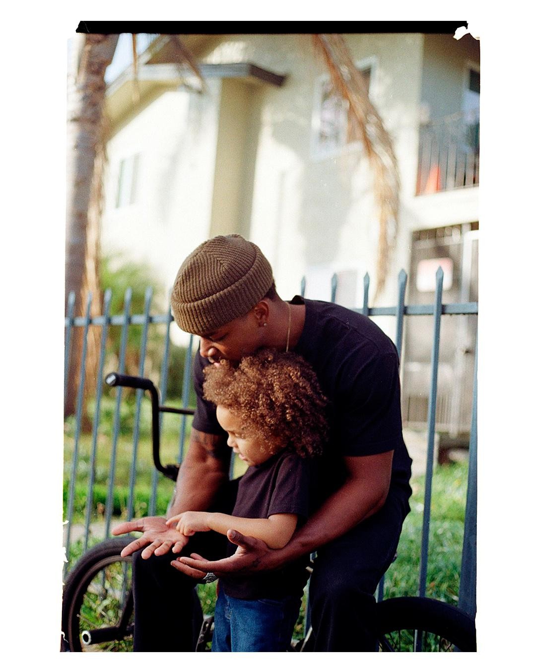 A man wearing a beanie holds a child with curly hair, who is pointing at the man's palm, in front of a metal fence and a building.