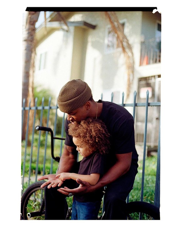 A man wearing a beanie holds a child with curly hair, who is pointing at the man's palm, in front of a metal fence and a building.