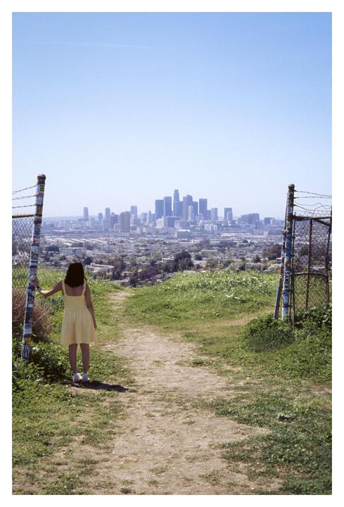 A woman in a yellow dress stands on a dirt path looking out at a city skyline.