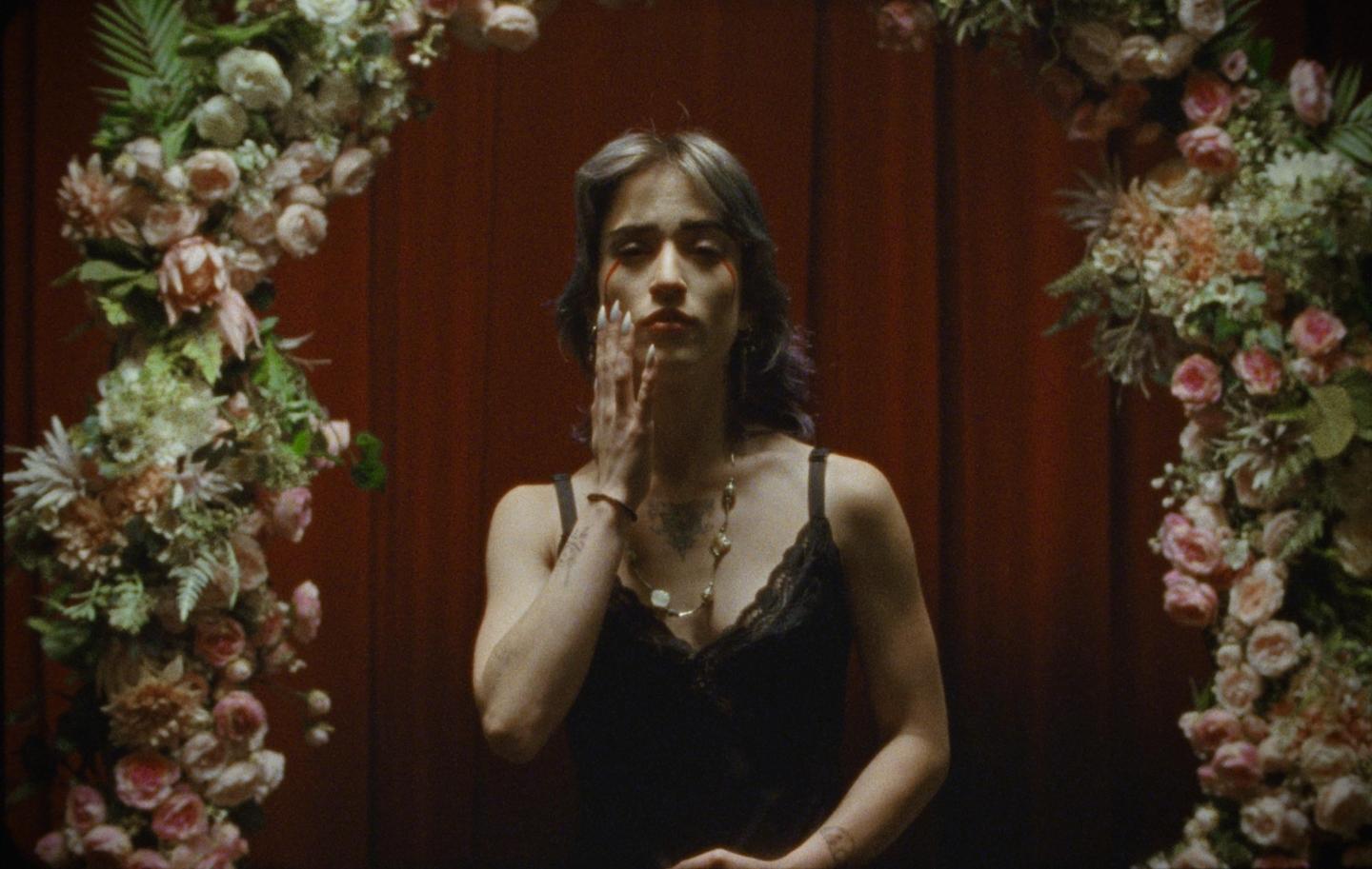 A woman with tears of blood on her face stands between two floral arrangements.