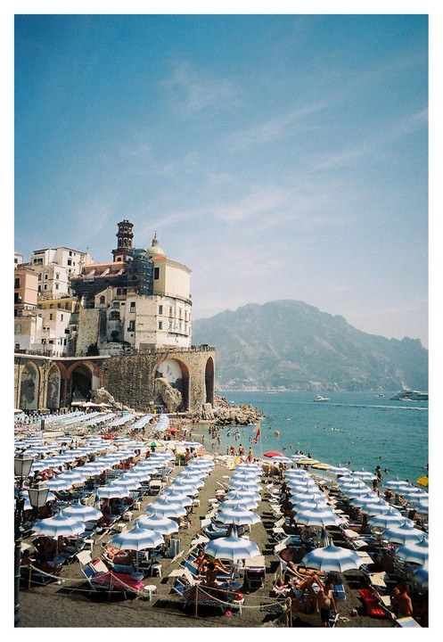A crowded beach with many striped umbrellas is next to a town built on a cliff overlooking the sea.