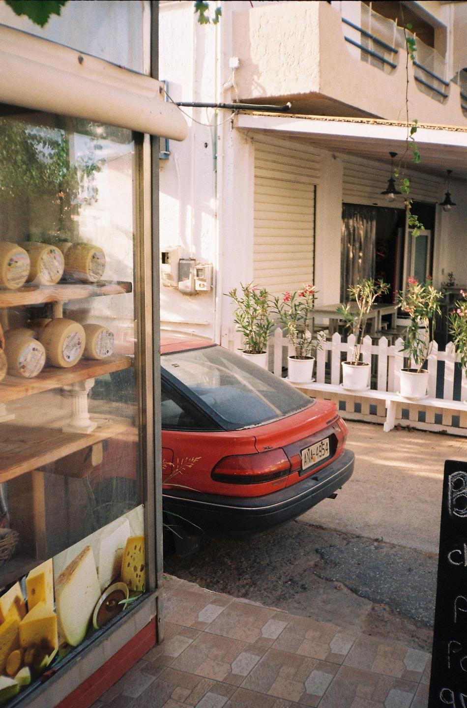 A red car is parked next to a shop window displaying cheese wheels and a white picket fence with potted plants.