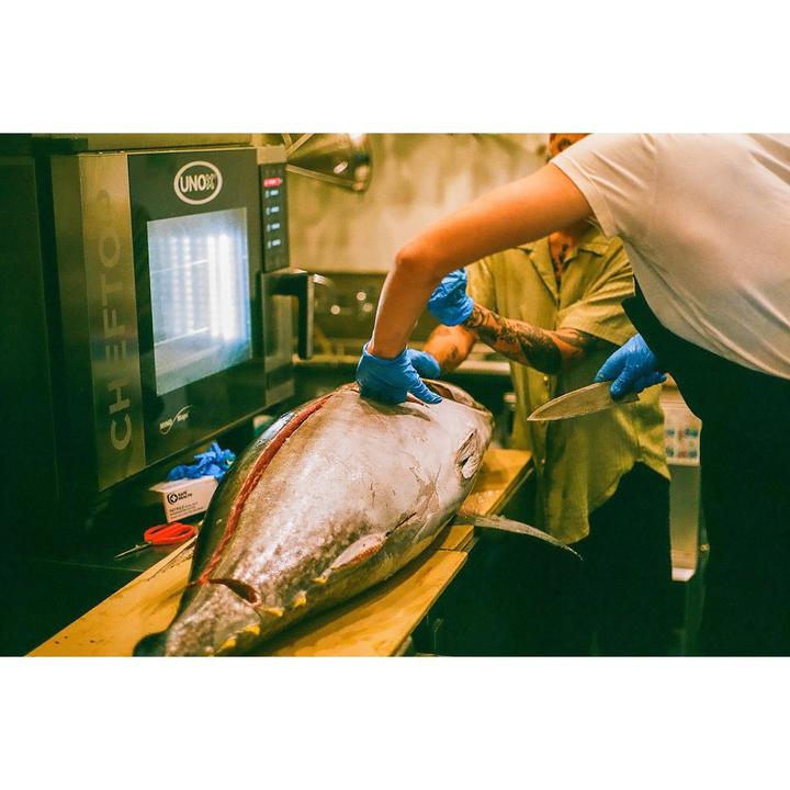 Two people wearing blue gloves are preparing a large tuna on a wooden board in a kitchen.