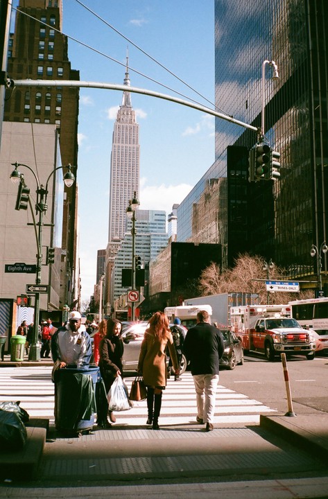 People cross a street in New York City with the Empire State Building visible in the background.