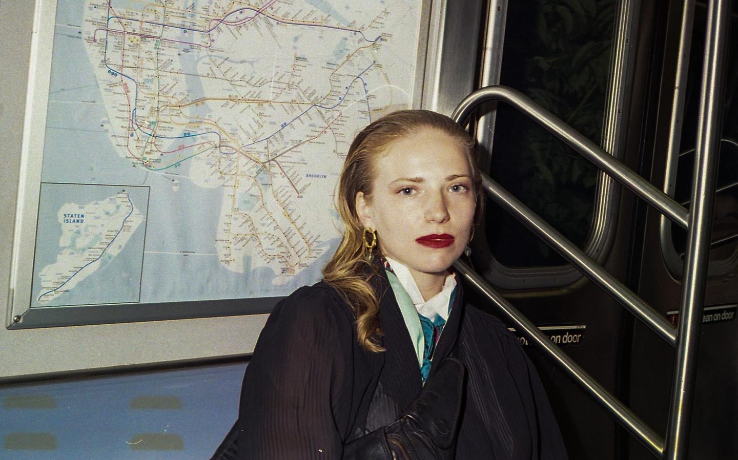 A woman with red lipstick and a scarf sits in a subway car with a map of New York City behind her.