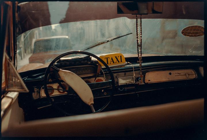 The interior of a taxi is visible through the windshield, with a rosary hanging from the rearview mirror.