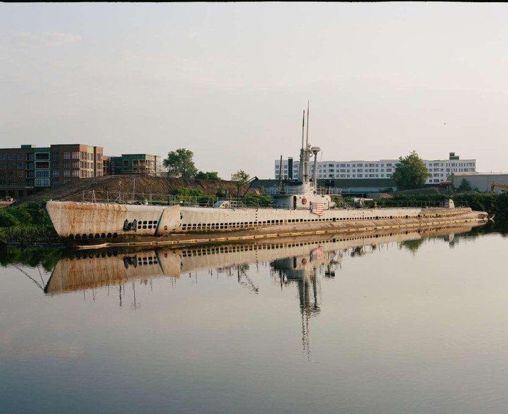 A large, rusty submarine is docked in a river with buildings in the background.