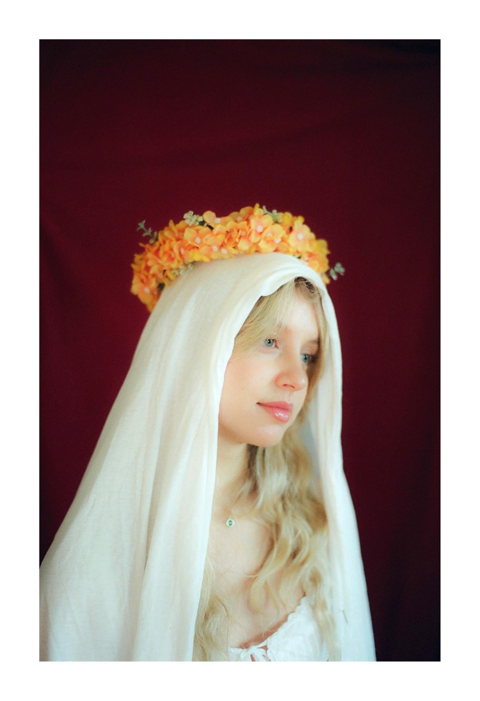 A woman with long blonde hair wears a white veil and a crown of orange flowers against a dark red background.