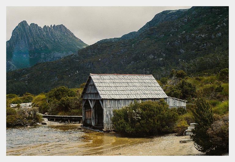 A wooden boathouse with a shingled roof sits on the shore of a lake with mountains in the background.