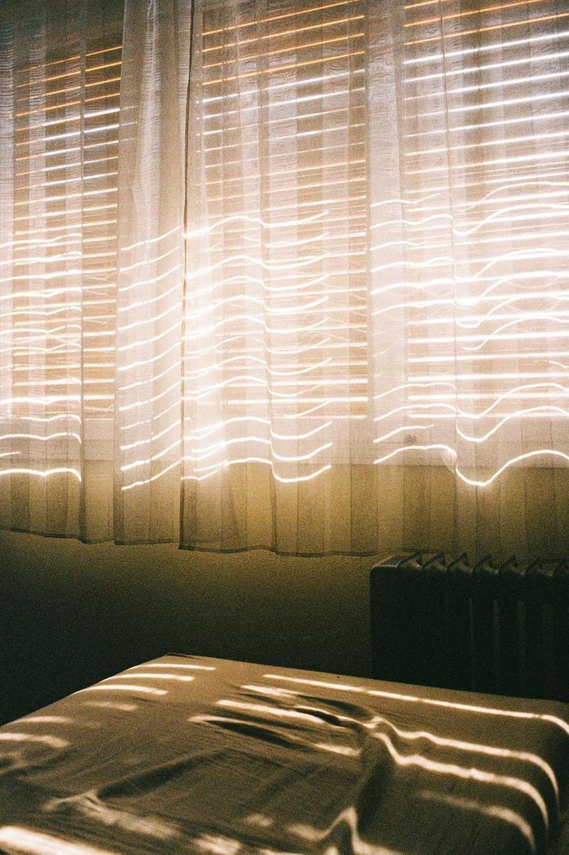 Sunlight streams through window blinds and curtains, casting patterns on a bed and wall.