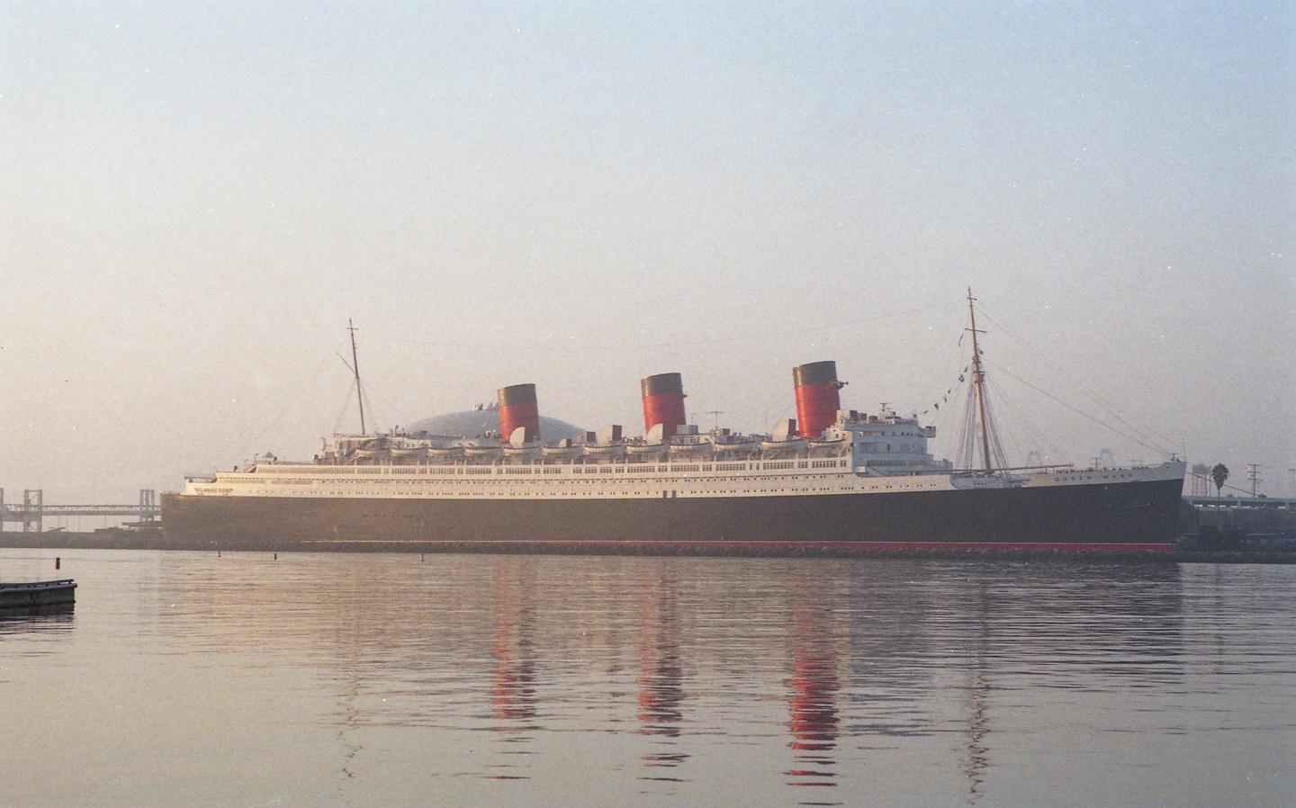 A large ship with three red and black smokestacks is docked in a harbor, with its reflection visible in the water.