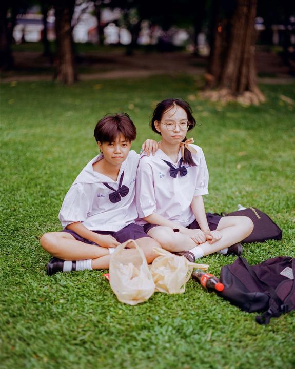 Two girls in school uniforms sit on a grassy lawn with plastic bags and a backpack.