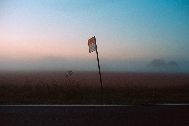 A bus stop sign stands beside a road and a field in a hazy landscape at dusk.