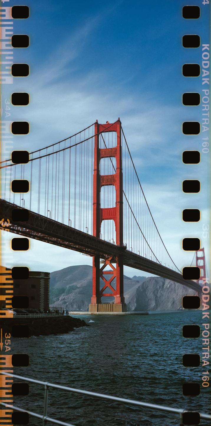 The Golden Gate Bridge spans across a body of water with mountains in the background under a blue sky.