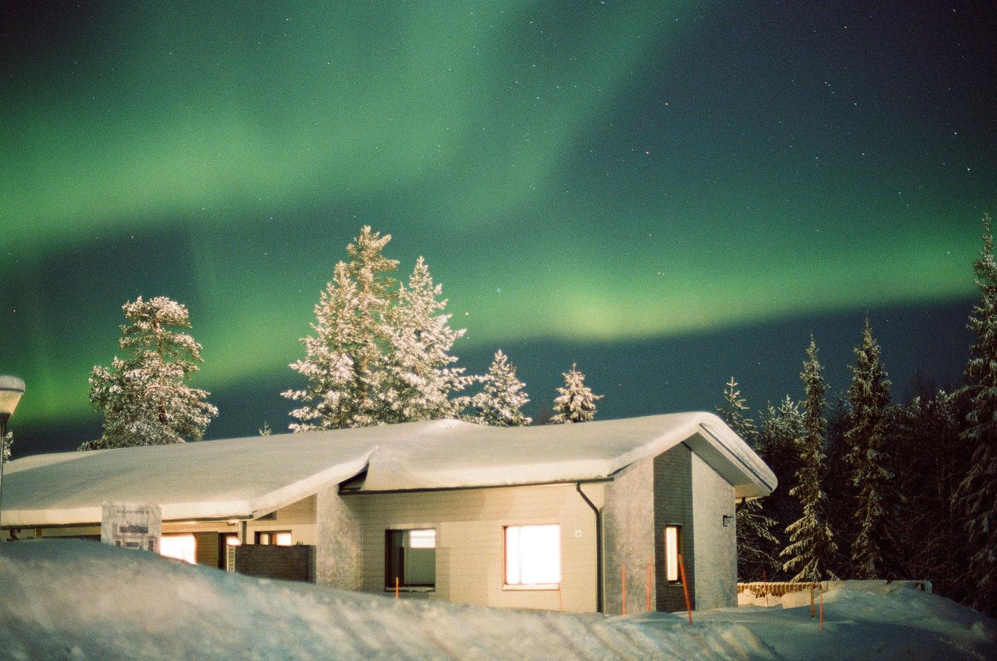 A snow-covered house with illuminated windows sits under a vibrant green aurora borealis and a starry night sky.
